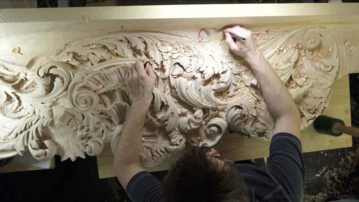 Seen from above, one hand marks red layout lines on the rough end of a long frieze already carved at the other end.