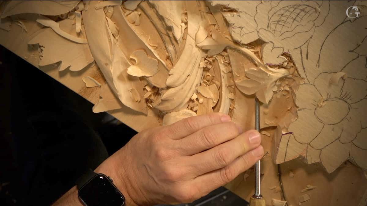 A close-up view of a person's hand skillfully carving details into a wooden surface. The carving tool is visible, creating intricate patterns and floral shapes. Shavings litter the workspace, highlighting the wood carving process. Perfect for showcasing wood carving classes and lessons!
