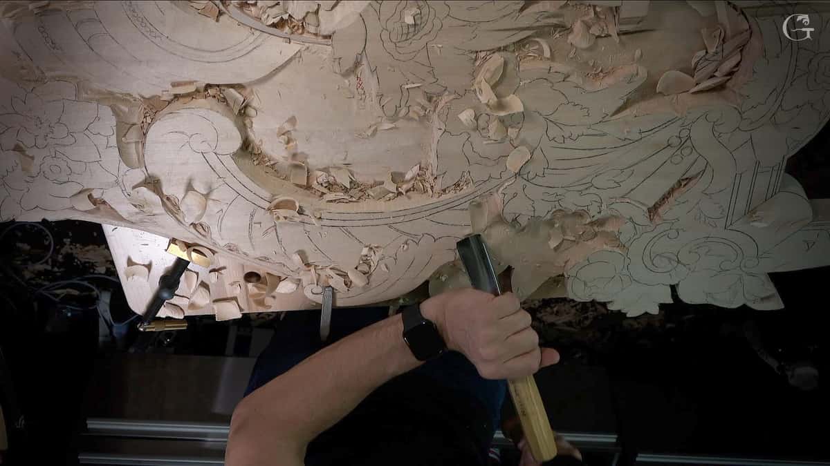 A close-up shot of a wood carving in progress shows intricate designs taking shape. The craftsman hammers a chisel into the wood surface, surrounded by shavings and curls from the work. This image captures the essence of wood carving lessons, showcasing skill and artistry that come with dedicated practice in wood carving classes.