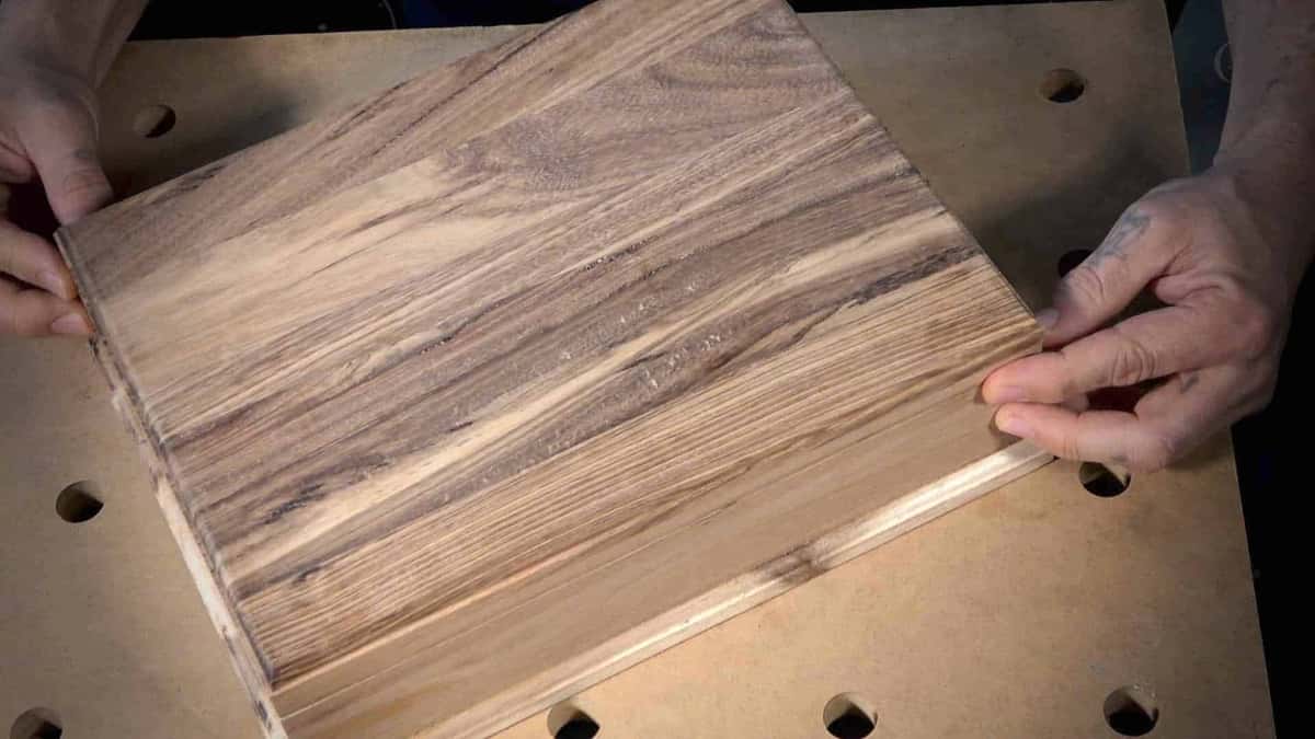 Hands tipping a planed dark streaked hardwood board to read its grain before carving