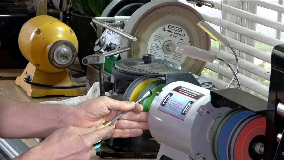 Carving tool held to a green buffing wheel beside a grinding wheel setup