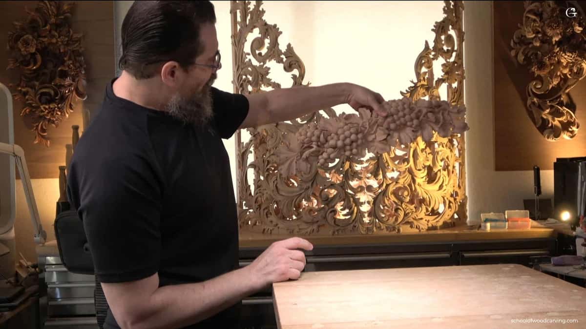 WOODCARVING LESSONS: Woodcarver Grabovetskiy with glasses and beard studies a grape wood carving. Another wood piece stands behind, softly lit. - Wood Carving tutorials