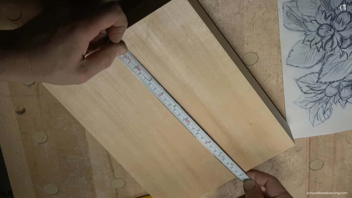 One hand holds a steel tape laid across a plain pale carving board, a pencilled hazelnut drawing right beside it