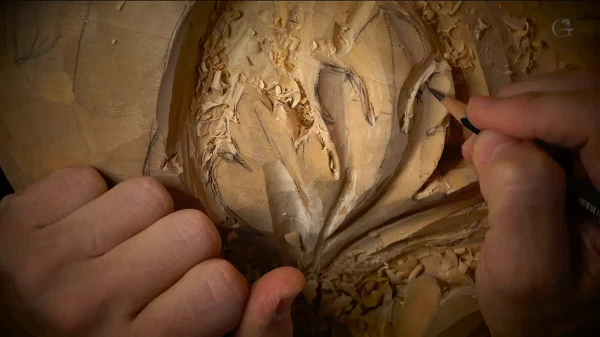 One hand holds the work while the other draws in pencil straight onto the half carved leaf, correcting the line.