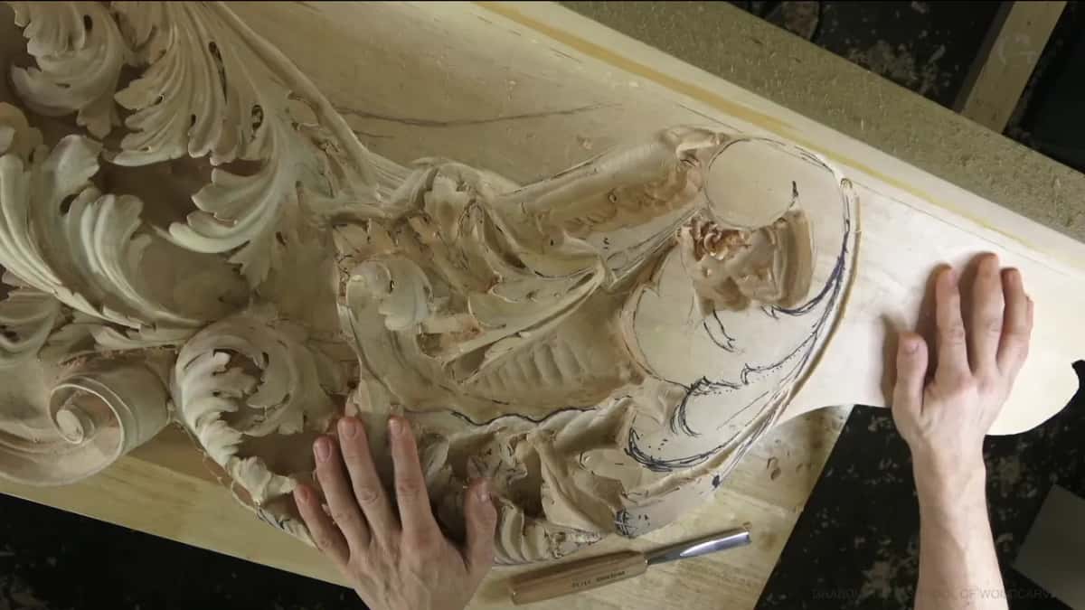Flat palms rest on a long carved panel where the next section is still only ink lines on bare wood.