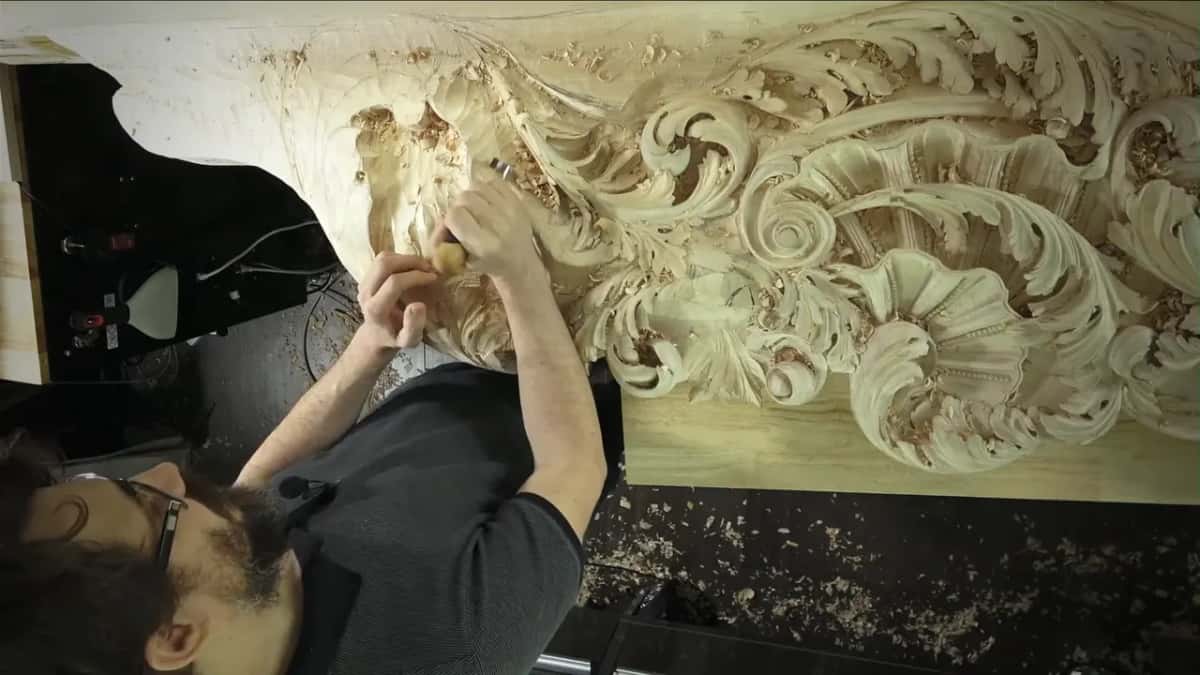 Looking down past his shoulder at a long panel of deep leaf carving, chips banked up on the bench below.