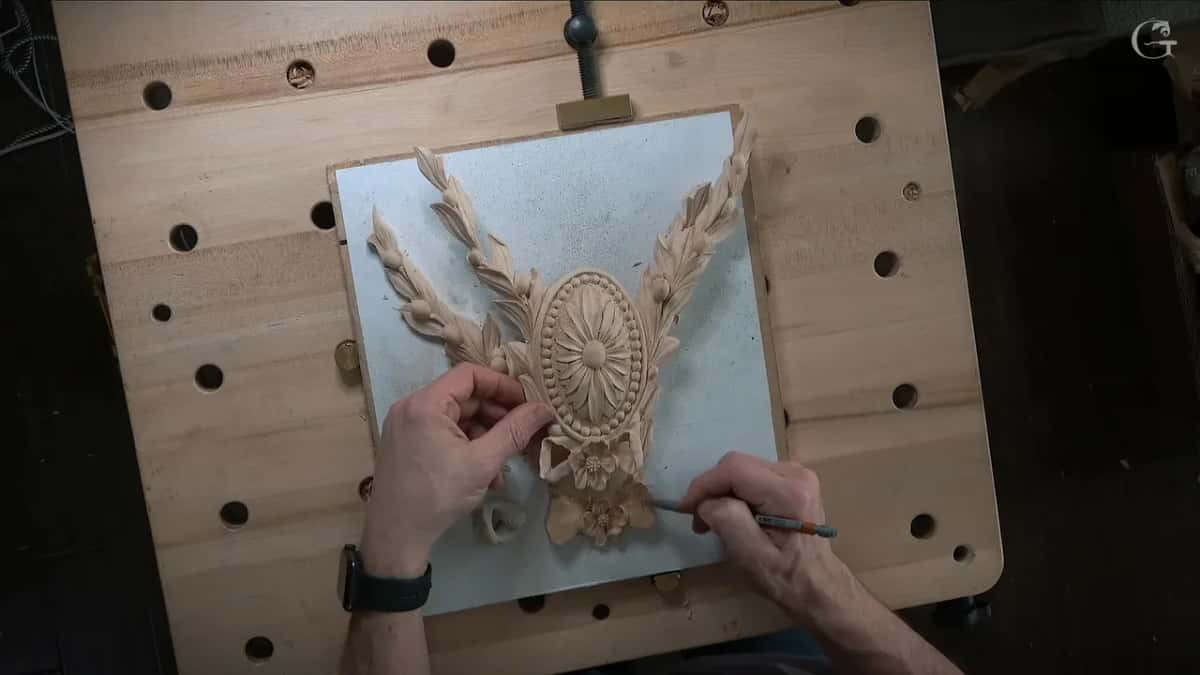 Both hands work at a rosette and laurel carving clamped to a bench board, a pencil marking its lower edge.