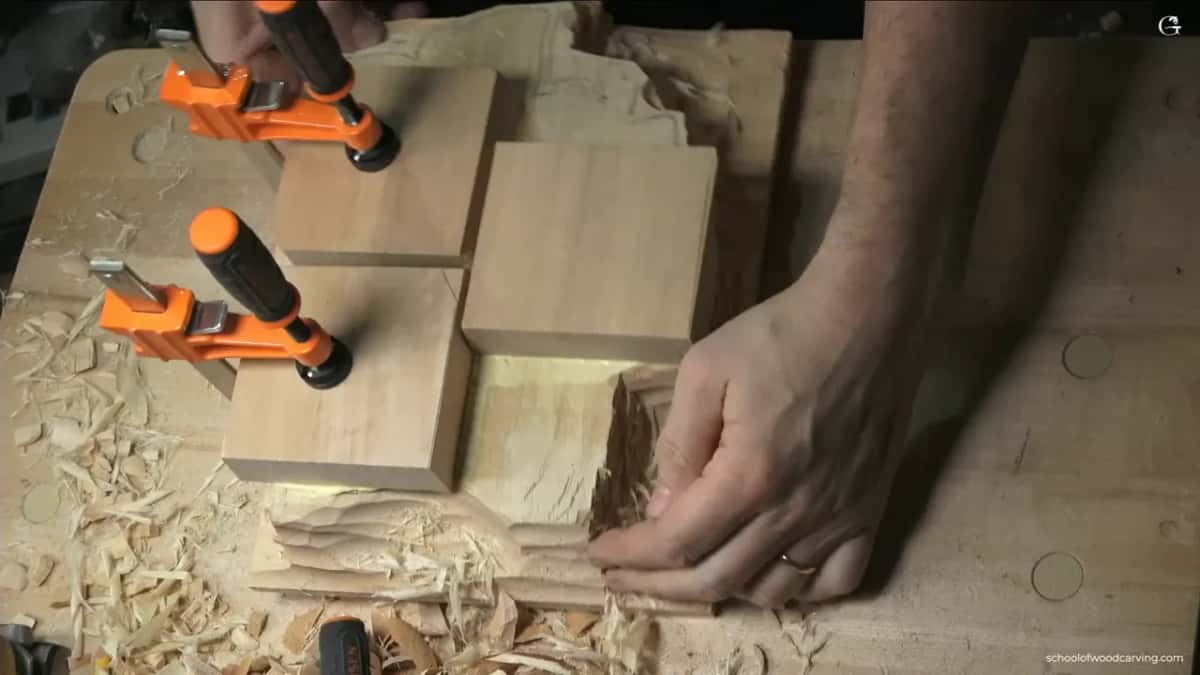 Two orange bar clamps hold a stack of blocks down while a hand steadies the carved panel underneath.