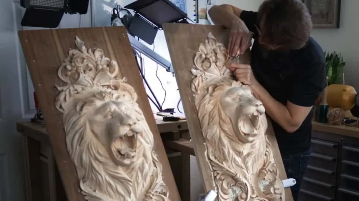 Lion heads carved on two tall panels stand at a bench, one of them still being worked on.