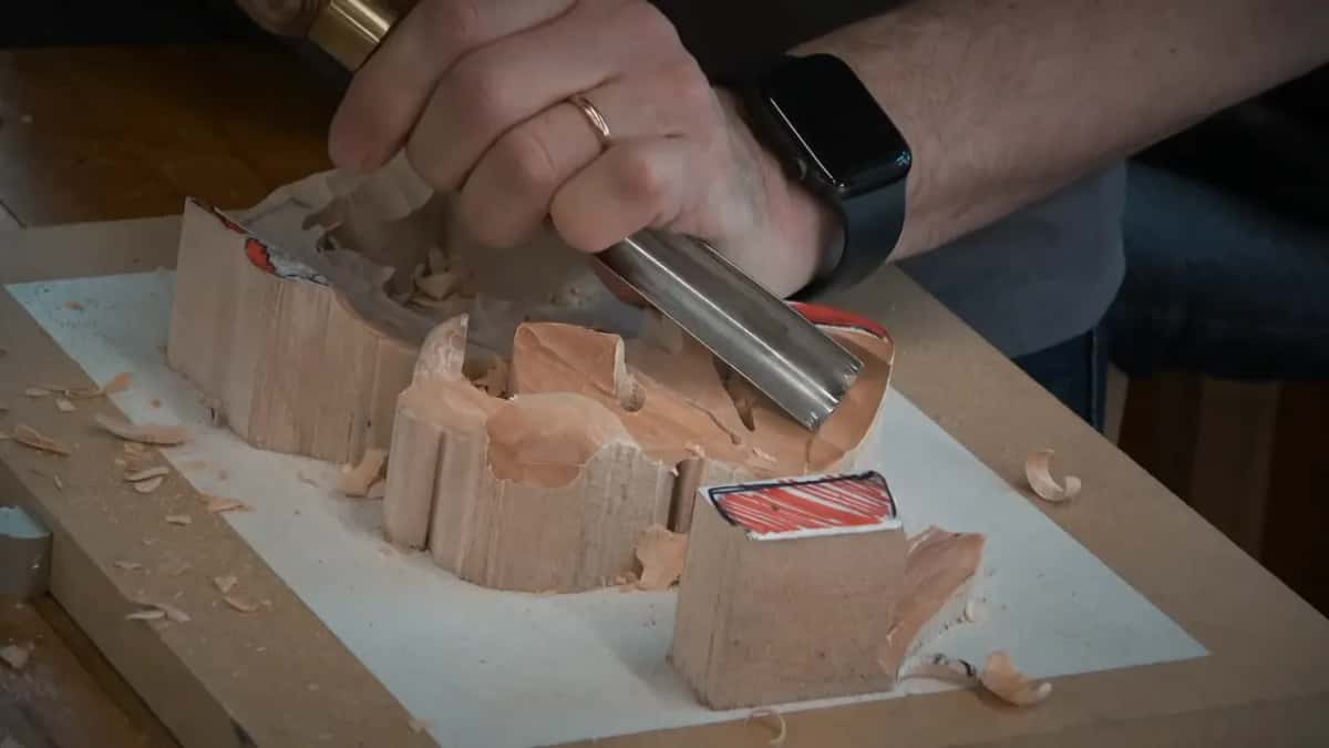 A wide curved tool carves the low ground between two standing blocks, with pale shavings scattered around.