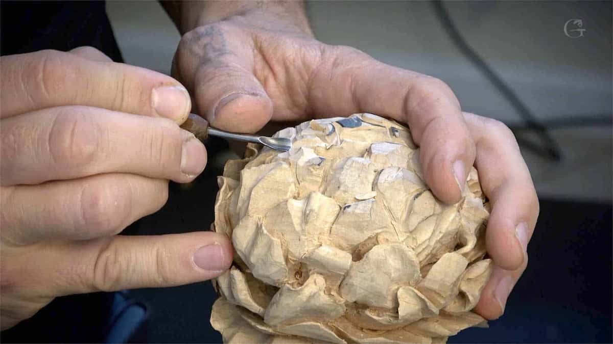Two hands cradle a round, deeply layered flower carved in pale wood while a small spoon gouge shapes one petal.