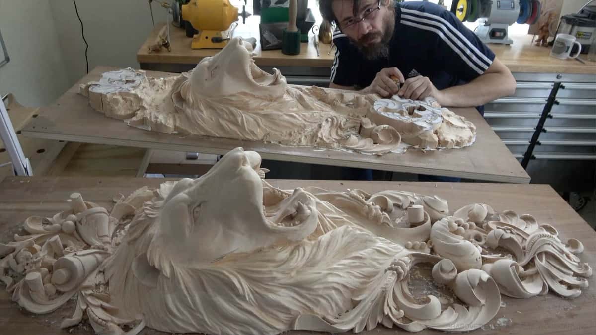 A wood carving enthusiast works on a detailed lion sculpture in a workshop filled with tools like chisels and a carving workbench. The intricate design features flowing mane and decorative elements, showcasing the beauty of craftsmanship. Perfect for those interested in wood carving classes or lessons!