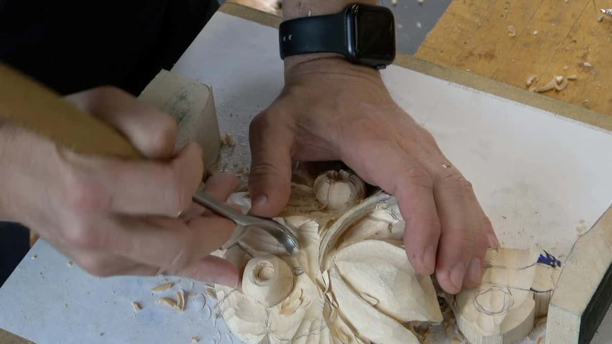 Hands modelling a carved rosette with a small bent gouge, fresh chips across the bench beside it