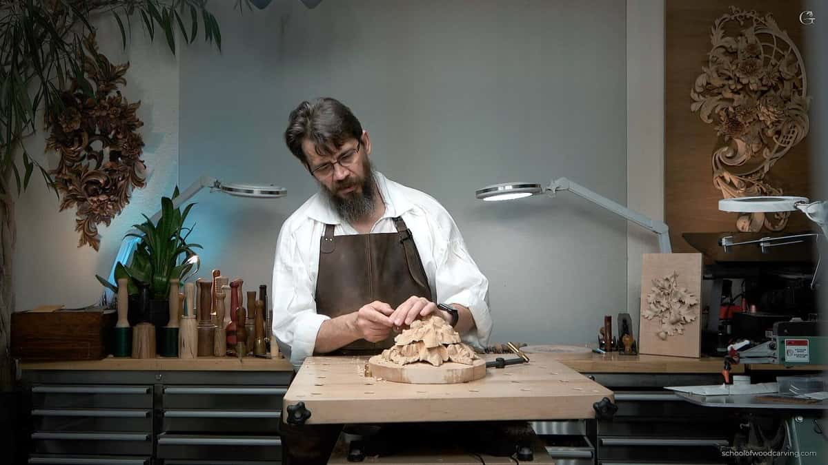 A woodcarver sits at a solid workbench holding carving tools, showing a proper woodcarving workbench setup