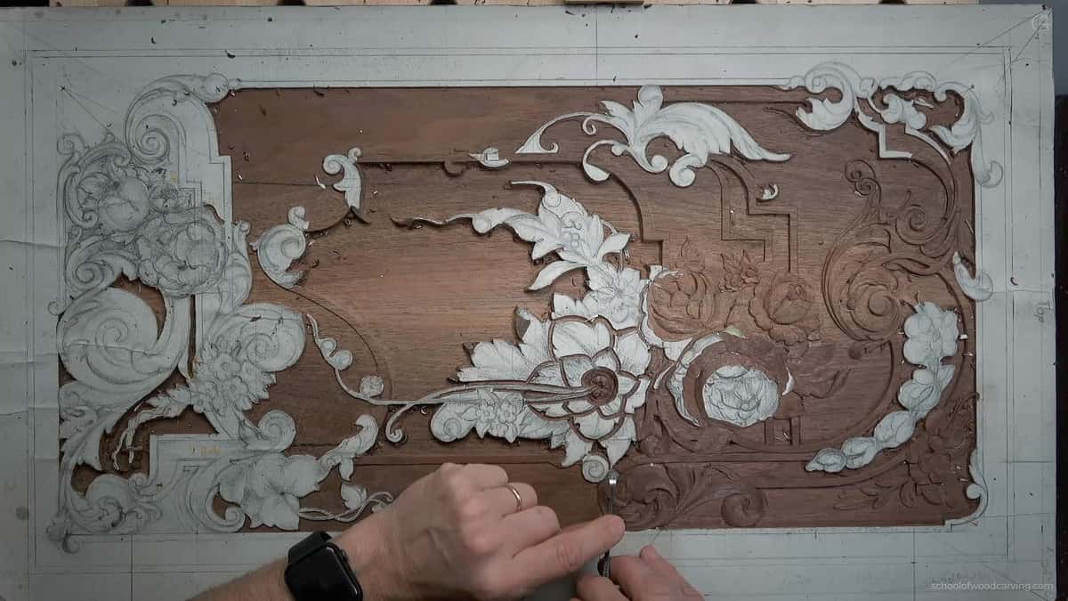 Alt text: A close-up of a wood carving project in progress, showcasing detailed floral designs etched into the surface. The artist's hands are seen delicately working, tools like chisels nearby, illustrating the intricate process of wood carving. Ideal for those interested in wood carving classes and lessons.