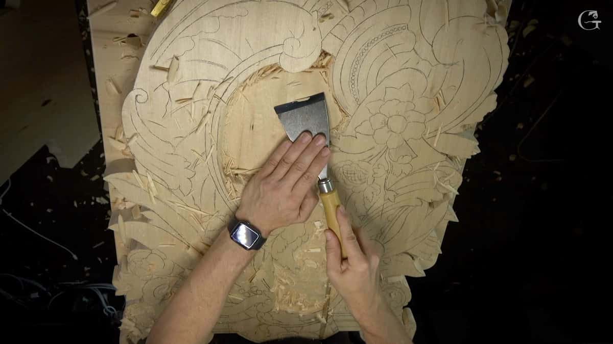A person is carving a detailed design into a piece of wood, using a large chisel with a wooden handle. Their hands are positioned carefully, guiding the tool along the outlines of flowers and leaves. The workspace shows shavings scattered around, hinting at the creative process of wood carving. This scene captures the essence of hands-on wood carving classes and lessons, where artisans bring their visions to life!