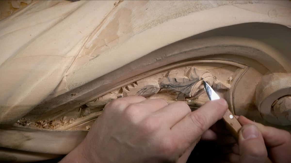 A knife point cutting a small leaf ornament inside a large curved basswood volute