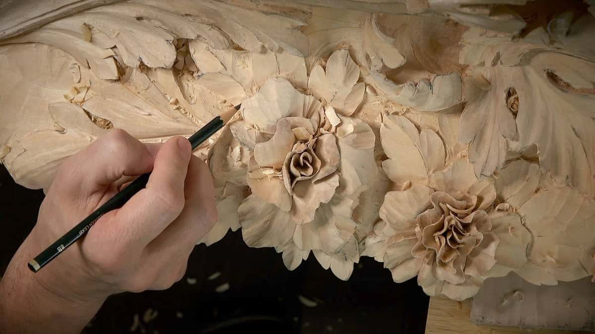 Hand holding a pencil over a high relief basswood spray of roses and leaves, fresh chips still lying on the carved surface