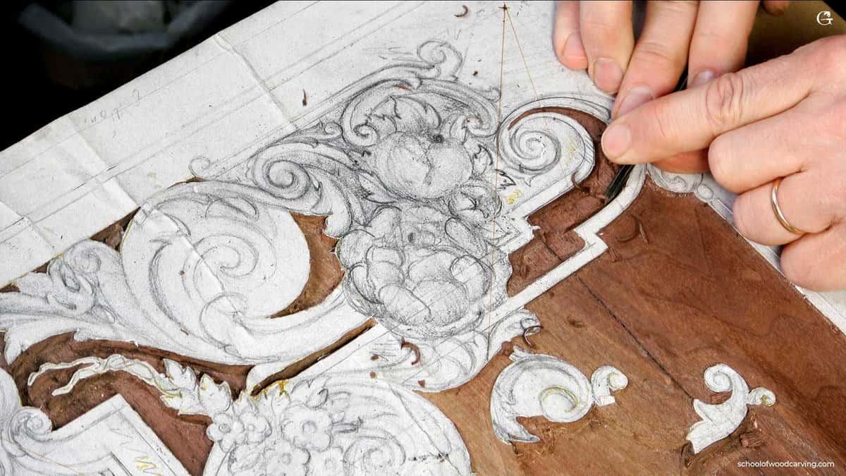 A craftsperson's hands carve a detailed floral design into a wooden panel using precision tools, with a sketch nearby for reference.