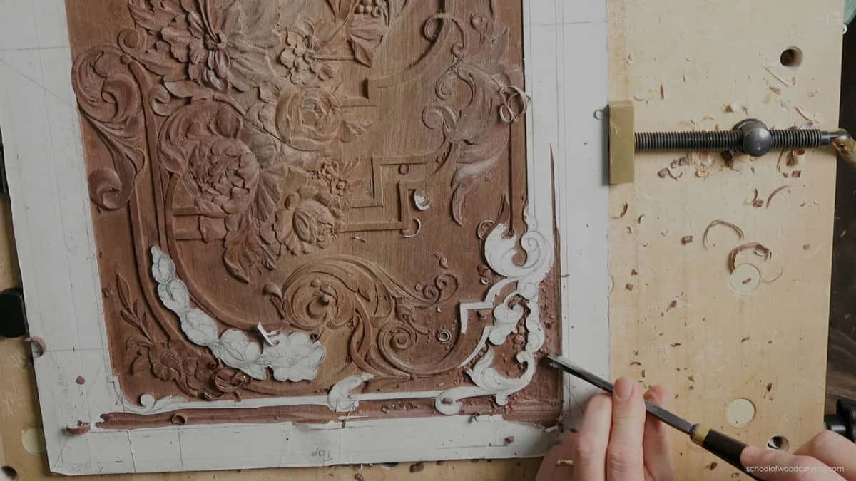 WOODCARVING LESSONS: A hand carves flowers and curly designs into a piece of wood using a carving tool. Sawdust is on the table, and you can see a metal clamp holding the wood. The carvings have flower shapes, leaves, and swirls. This photo shows Woodcarver Grabovetskiy working during his woodcarving lesson or tutorial. - Wood Carving tutorials