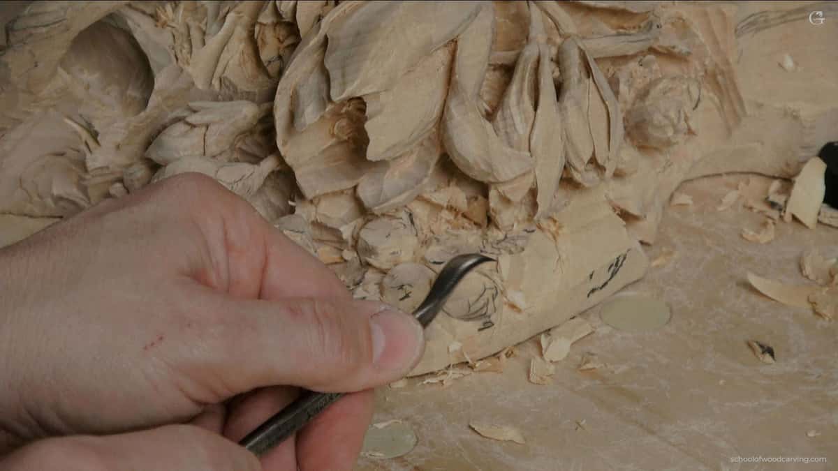 WOODCARVING LESSONS: A close-up photo shows Woodcarver Grabovetskiy holding a carving tool. He carefully carves detailed flower and leaf shapes into wood. You can see wood shavings and carved patterns on the wood's surface. The table has small pieces of wood scattered around. Learn how to carve beautiful designs with expert Woodcarver Grabovetskiy in our easy-to-follow woodcarving lessons or course. See the tools, clear carving steps, and tips for beginners in every tutorial picture. - Wood Carving tutorials