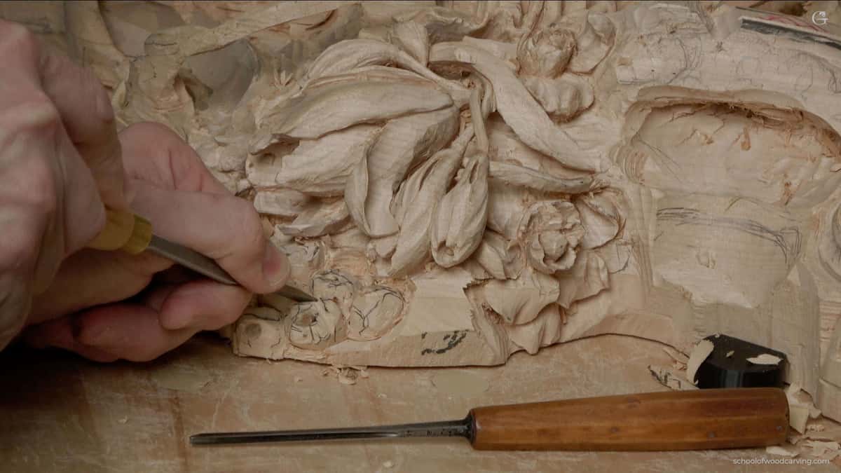 WOODCARVING LESSONS: Close-up photo of Woodcarver Grabovetskiy's hand using a chisel for woodcarving. He is carving beautiful flower patterns into light wood. Another chisel is on the table next to the wood. This picture shows detailed and careful woodcarving work during a lesson or tutorial with master Grabovetskiy. Learn how to carve floral designs in wood step by step in this course. - Wood Carving tutorials