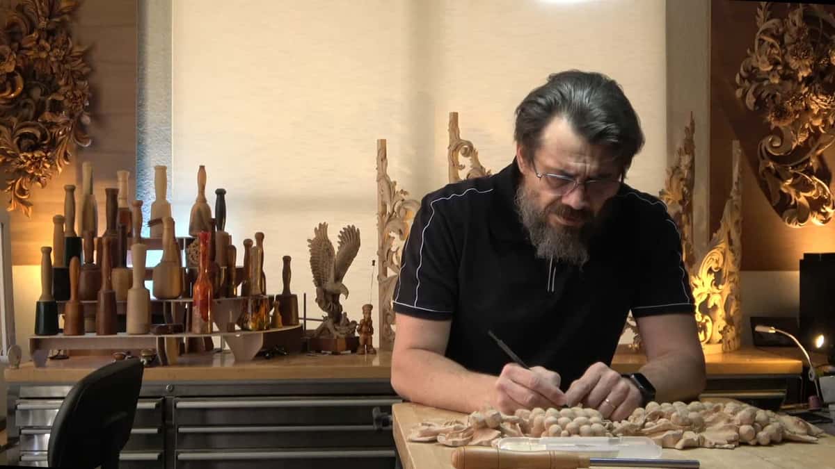 WOODCARVING LESSONS: Woodcarver Grabovetskiy, with glasses and beard, carves wood at a table with tools and sculptures, perfect for woodcarving lessons. - Wood Carving tutorials