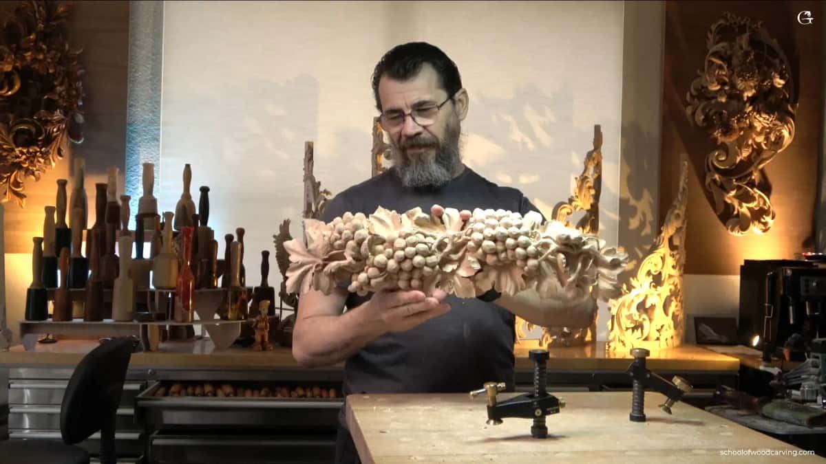 WOODCARVING LESSONS: Woodcarver Grabovetskiy in his workshop holds a carved wood grape leaf ornament. Wood tools and panels are behind him, warmly lit. - Wood Carving tutorials