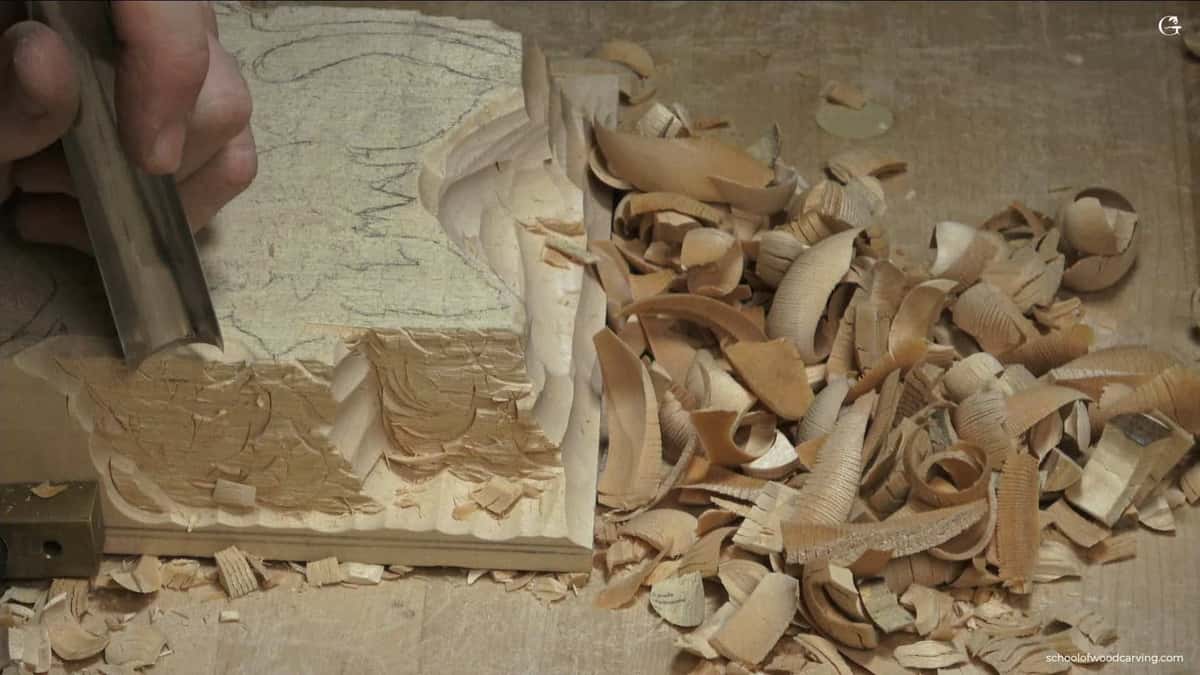 WOODCARVING LESSONS: Woodcarver Grabovetskiy uses a chisel to carve wood. Curled wood shavings and chips surround the textured wooden block. - Wood Carving tutorials