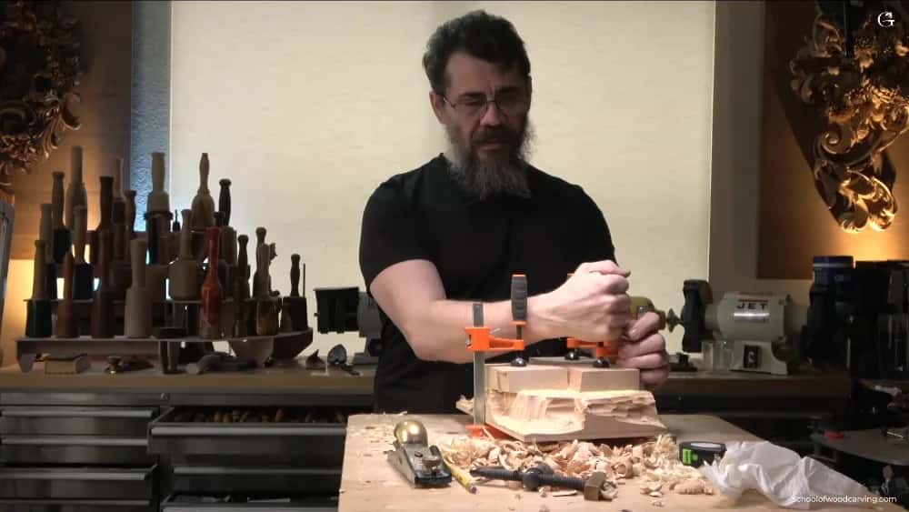 WOODCARVING LESSONS: Woodcarver Grabovetskiy clamps wood at a workbench with tools and shavings. Hazelnut carvings and spindles shown behind him. - Wood Carving tutorials
