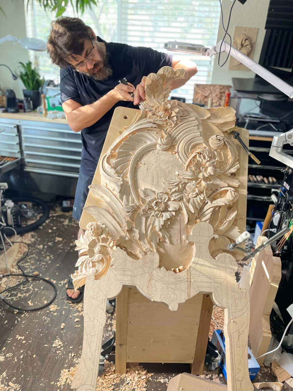 A bearded carver shapes a large floral relief carving with a chisel in a workshop