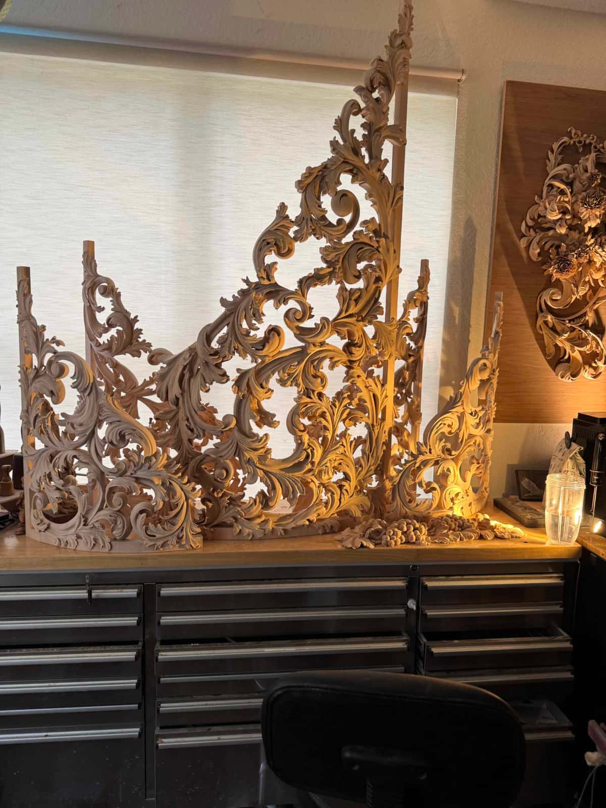 A large acanthus leaf carving with scrolling vines and grape clusters rests on a workbench in warm light