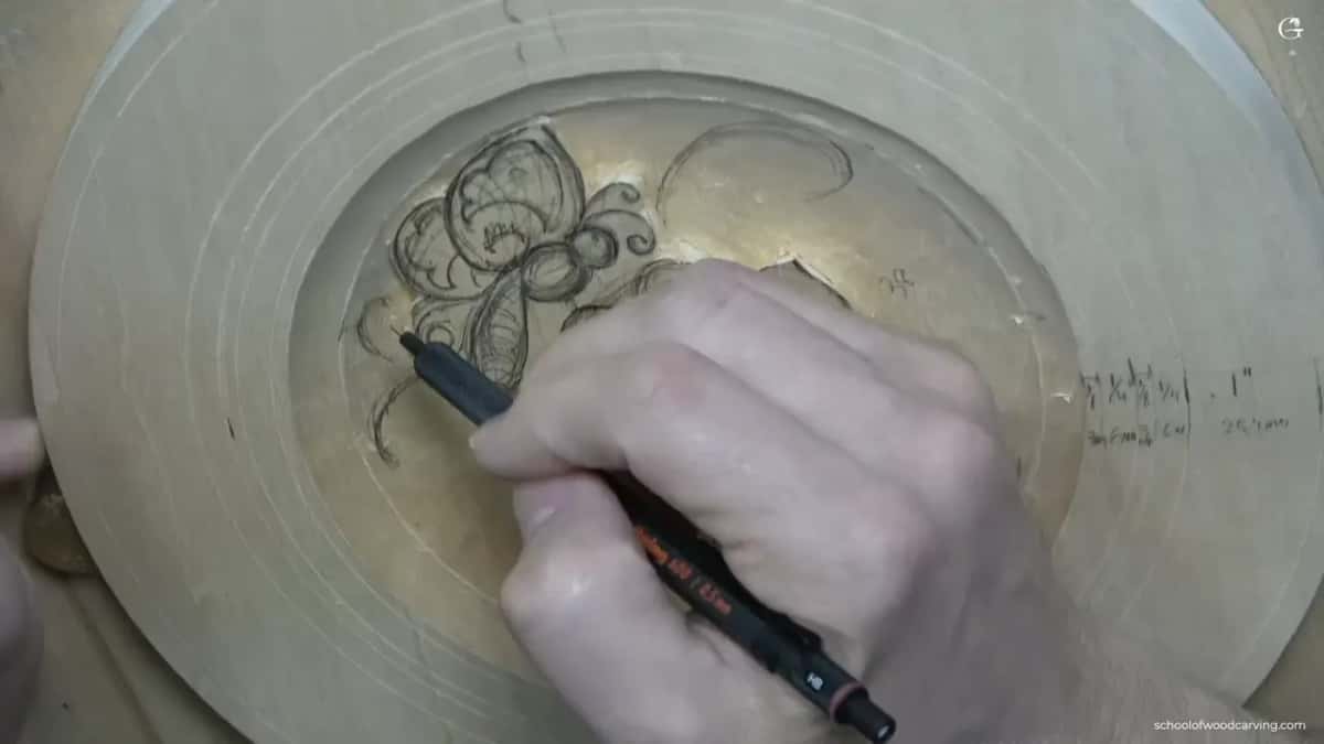 A hand draws a flower in pencil inside the hollow of a round pale blank, sizes written on the rim.