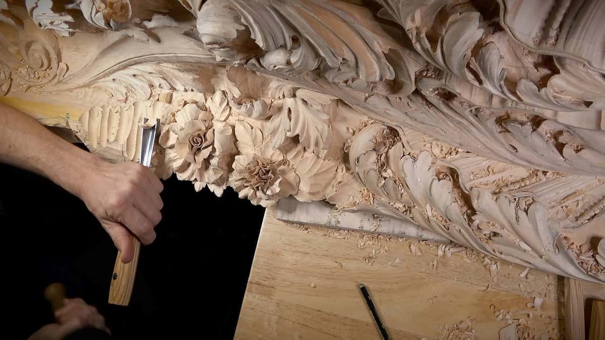 A carver guides a gouge along a large floral relief carving with roses, leaves, and scattered wood shavings