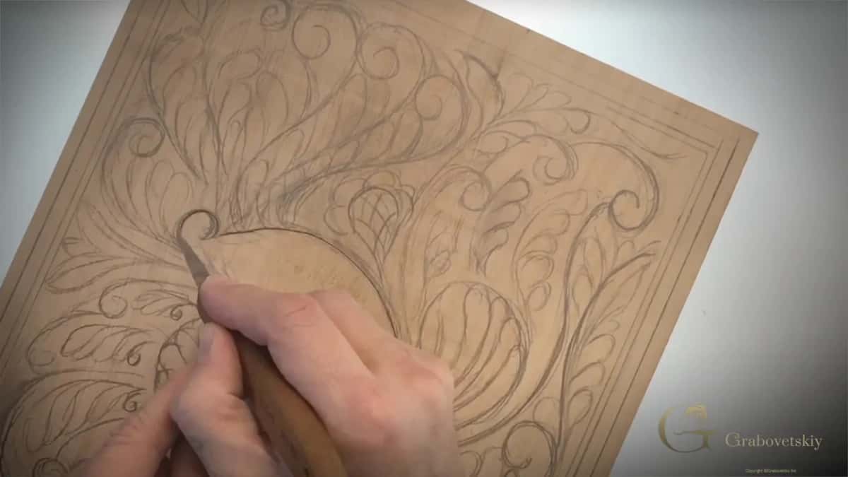 Knife cutting around one large drawn curl on a pale board covered in a pencilled scrolling design