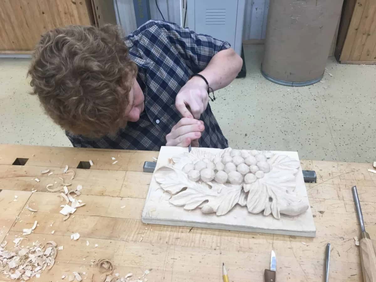 Hands carving a cluster of berries into basswood, chips scattered right across the bench