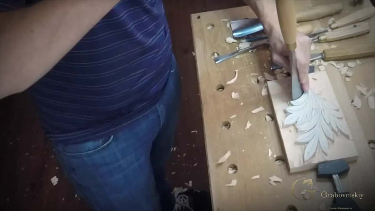 A gouge pressed by hand into a carved acanthus leaf, two mallets lying idle on the bench