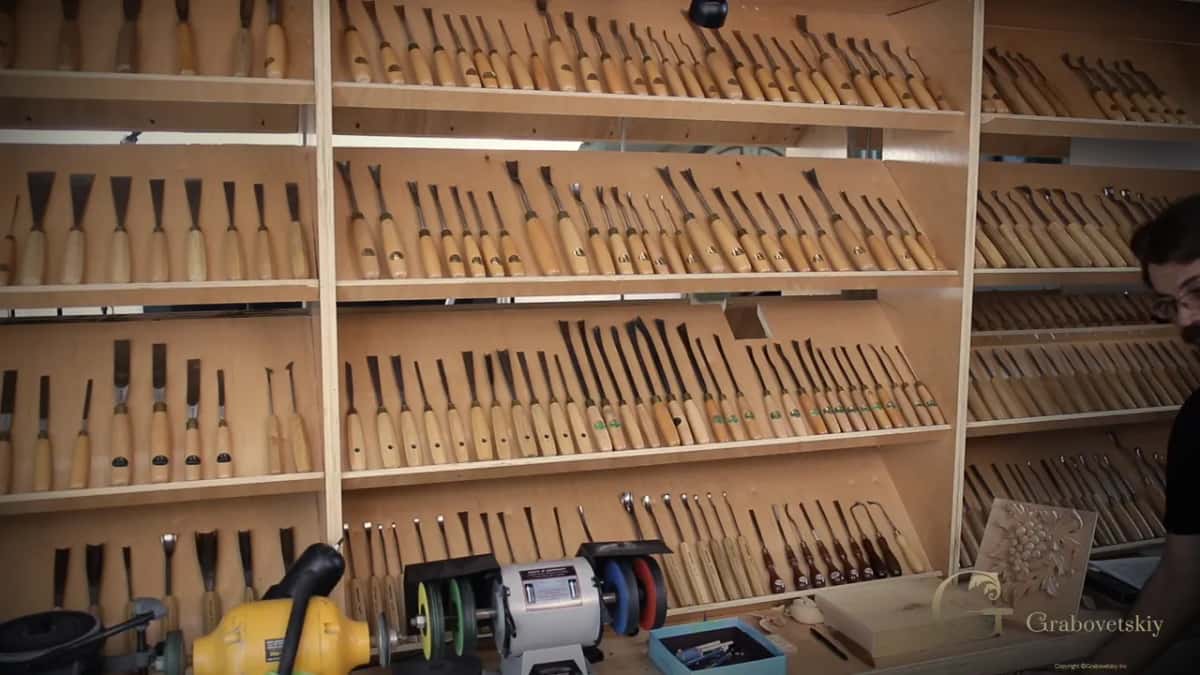 Four shelves of racked carving gouges and chisels above a bench grinder in the shop