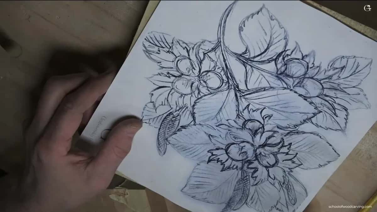 A hand holding the pencil drawing of hazelnuts and leaves, before a single chip is carved