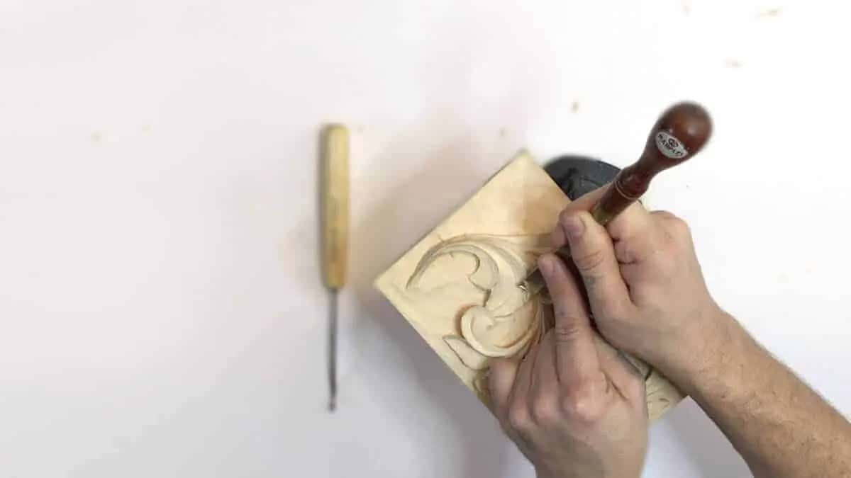One small gouge resting on a white bench while a second tool works a carved acanthus block