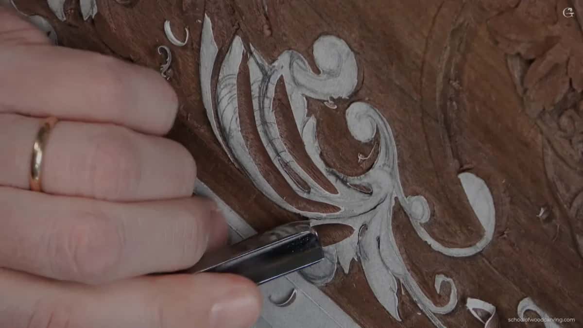 A bright gouge blade set against a drawn acanthus on dark reddish wood, hand steady at the left.