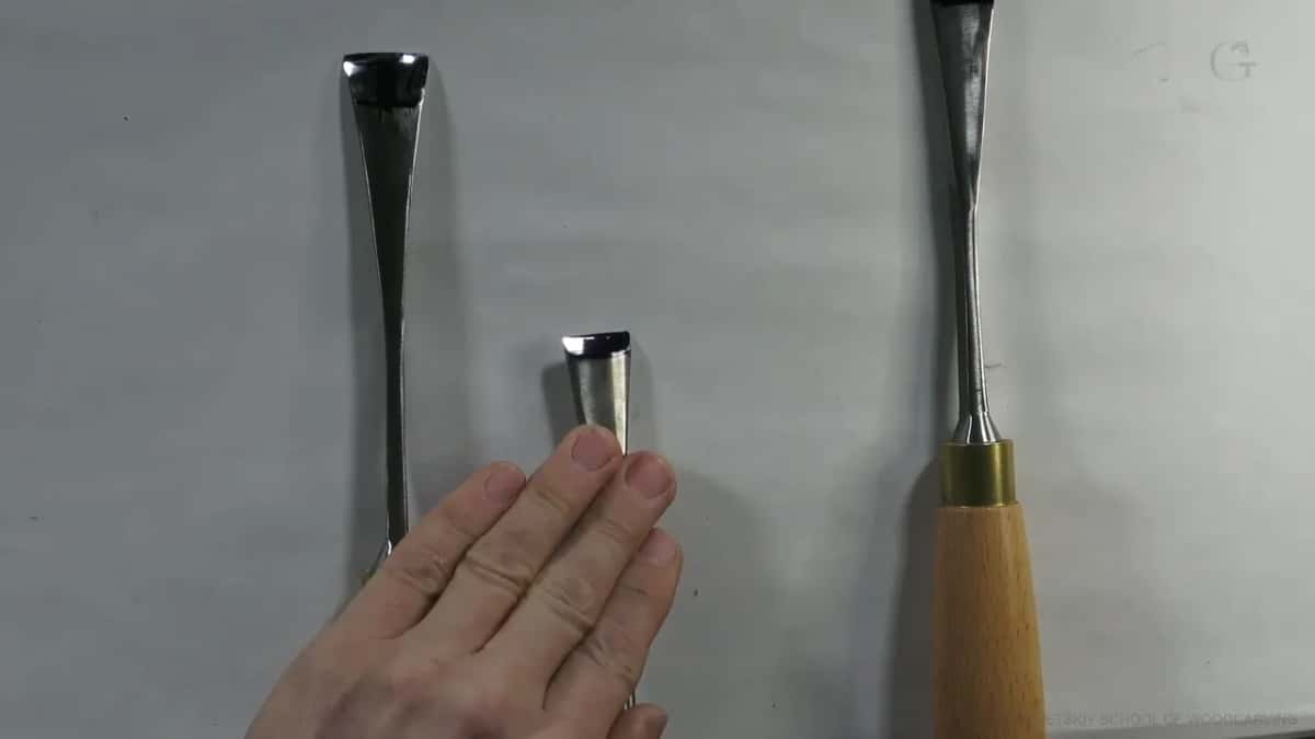 Three carving gouges laid on white, bevels toward the camera, a hand pointing at the middle blade.