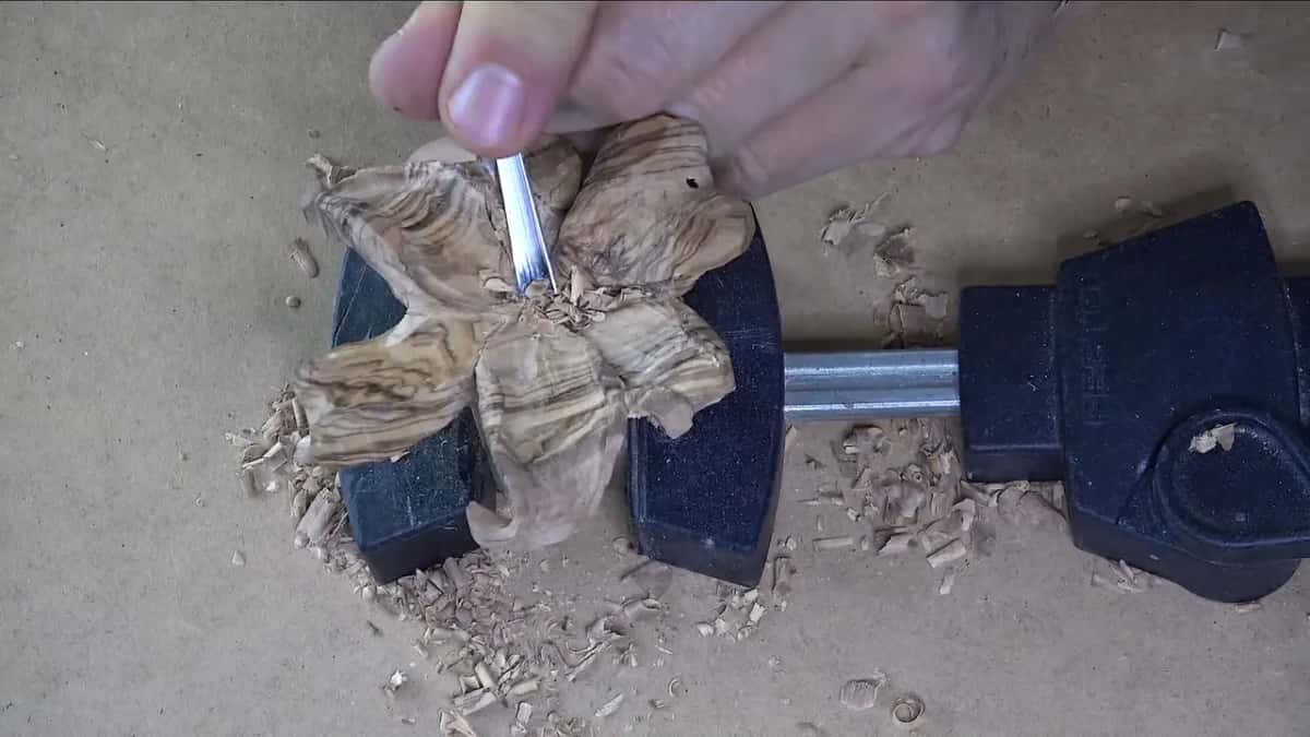A small olive flower gripped in a black clamp while a fine tool shapes its centre, chips all around.