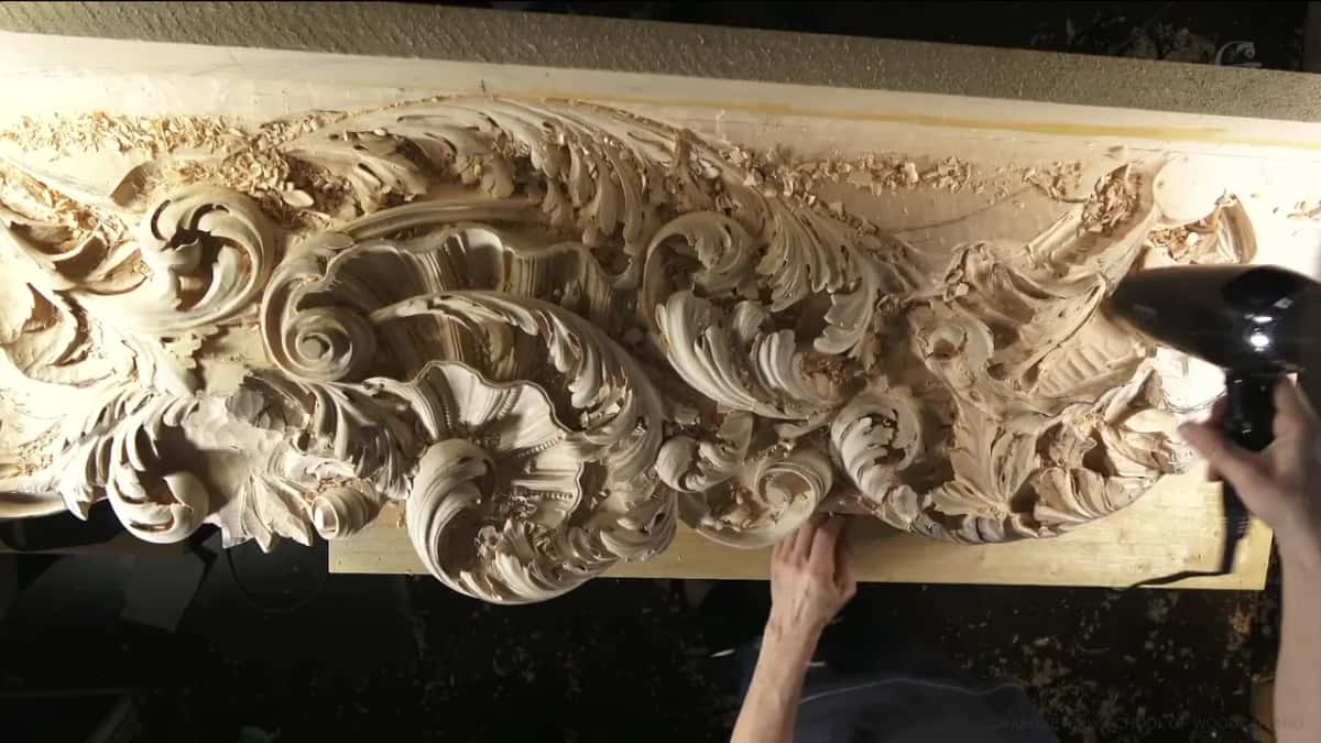 A black hair dryer is held over a long acanthus frieze on the bench, one hand steadying the carving.