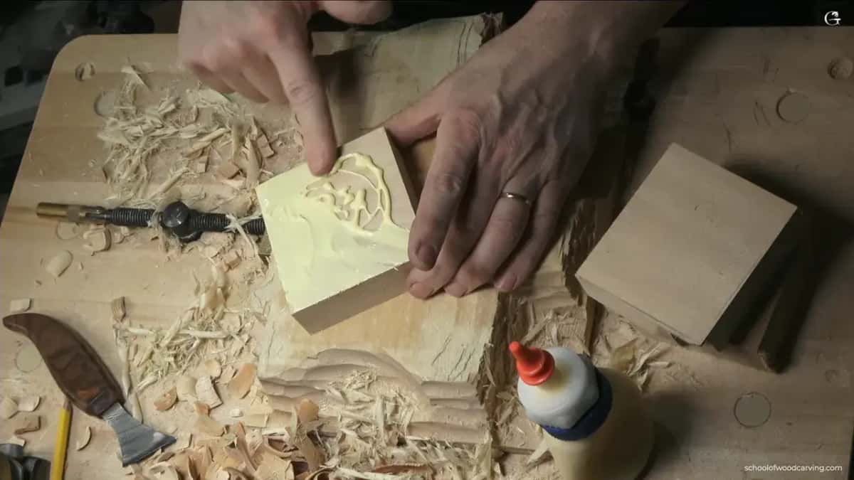 Both hands pressing a small pale carving blank down onto a backing block, its underside spread with yellow glue