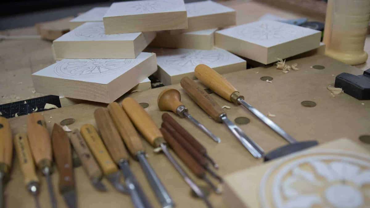 Row of carving gouges and knives laid out on a bench beside pale blanks marked with pencil rosette layouts