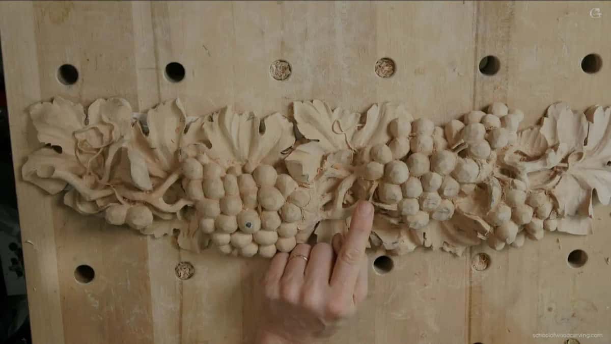 Bench top shows through the gaps between the leaves and stems of a finished grape garland, and a hand rests below the carving to give the scale of the openings that go clean through the board