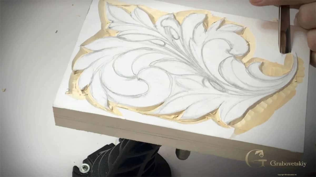A bare hand pushing a gouge to clear the background around a drawn acanthus panel