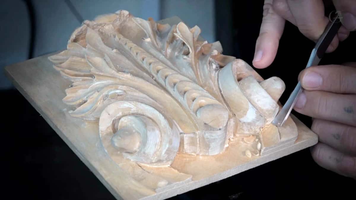 Carved acanthus panel held unclamped in two hands while a narrow tool works its right edge