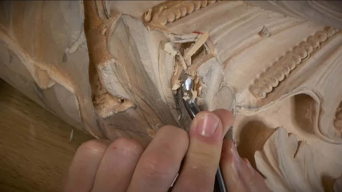 A small gouge worked into a tight corner of a deep leaf carving, one sliver of fiber lifting at the blade.